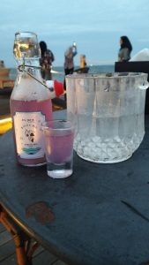 A bottle of purple beverage with a small glass and an ice bucket on a table, overlooking the sea with visitors relaxing in the background. 桌上摆放着一瓶紫色饮品、小玻璃杯和冰桶，背景是美丽的海景以及正在悠闲放松的游客。